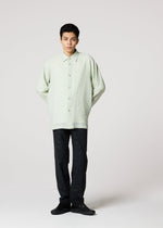 Against a light gray background, a person wears IM MEN’s STRIPE LINEN SHRINK shirt in light green with loose dark pants and black shoes, hands behind their back and a neutral expression.