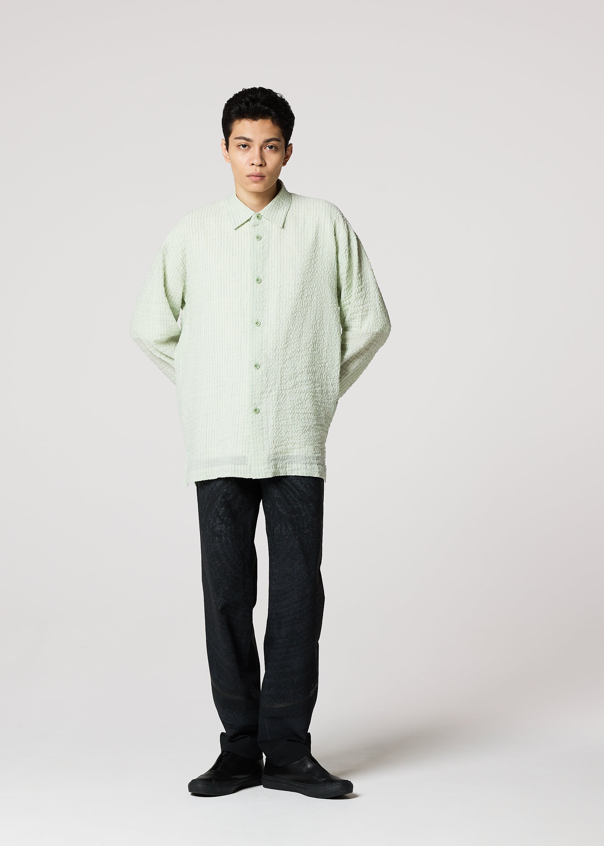 Zoomed view Against a light gray background, a person wears IM MEN’s STRIPE LINEN SHRINK shirt in light green with loose dark pants and black shoes, hands behind their back and a neutral expression.