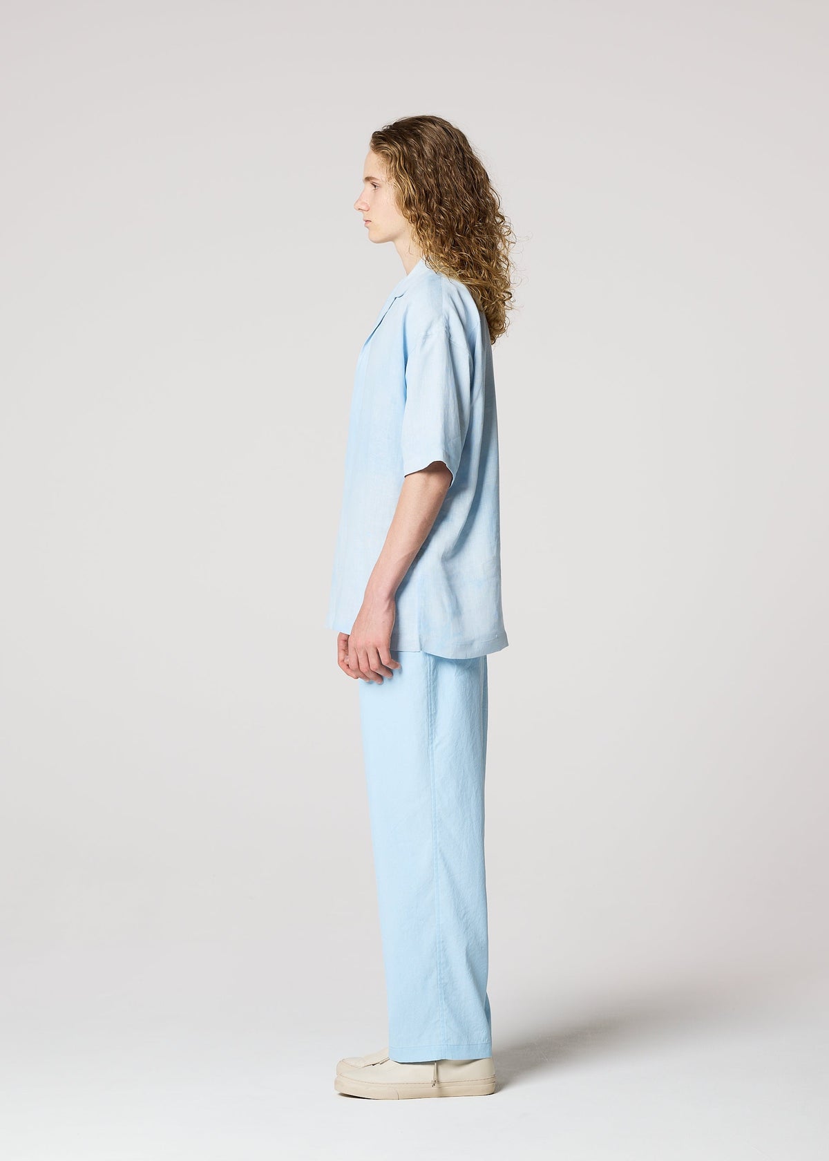 A person with long curly hair stands in profile against a plain background, wearing IM MEN’s FLOATING LINEN loose-fitting sheer shirt and matching pants, paired with beige shoes. view 7 of 9