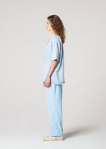 A person with long curly hair stands in profile against a plain background, wearing IM MEN’s FLOATING LINEN loose-fitting sheer shirt and matching pants, paired with beige shoes.