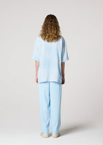A person with curly hair faces away from the camera, wearing IM MEN's FLOATING LINEN light blue blended shirt and matching pants with beige shoes, set against a plain light background.