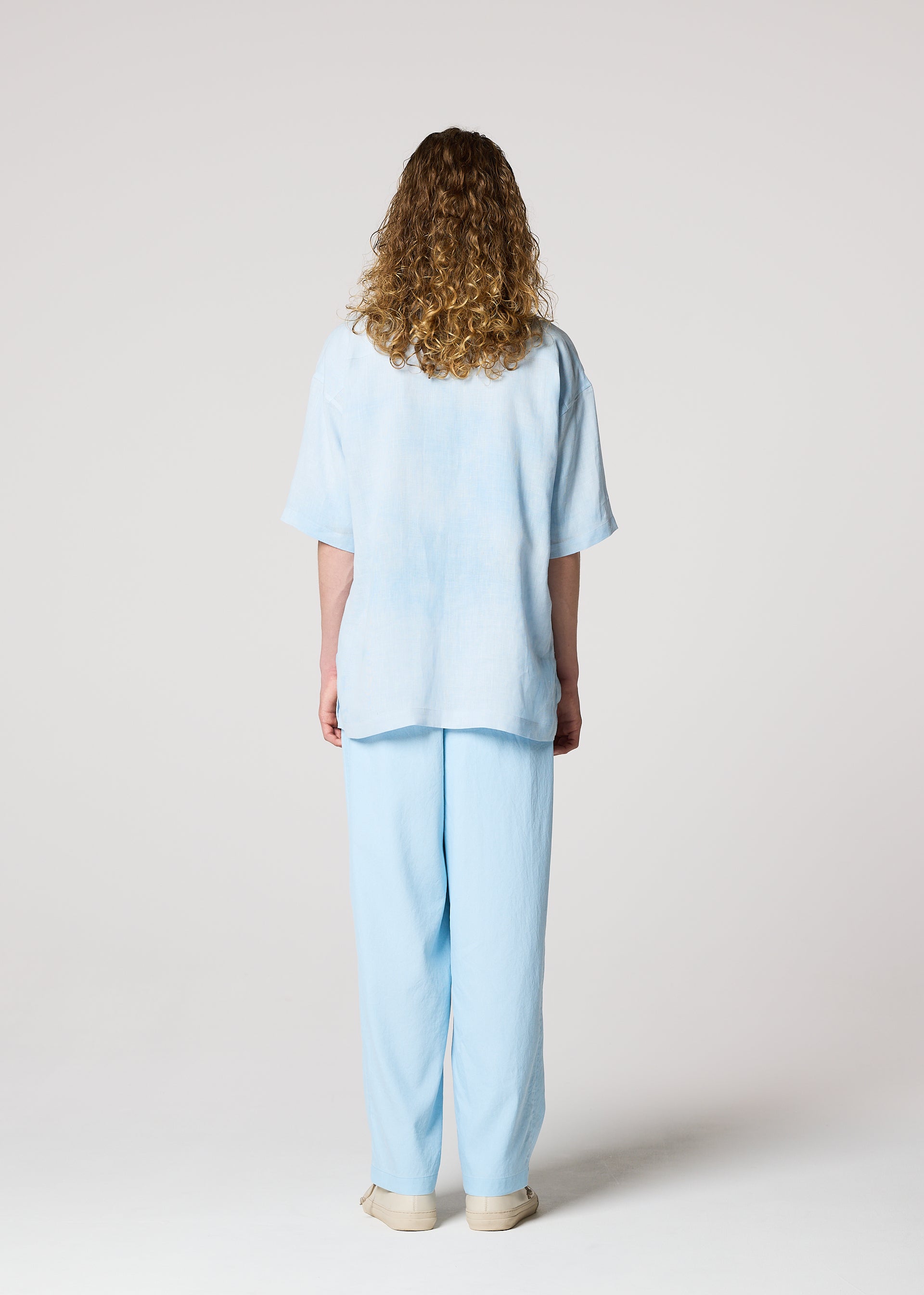 Zoomed view A person with curly hair faces away from the camera, wearing IM MEN's FLOATING LINEN light blue blended shirt and matching pants with beige shoes, set against a plain light background.