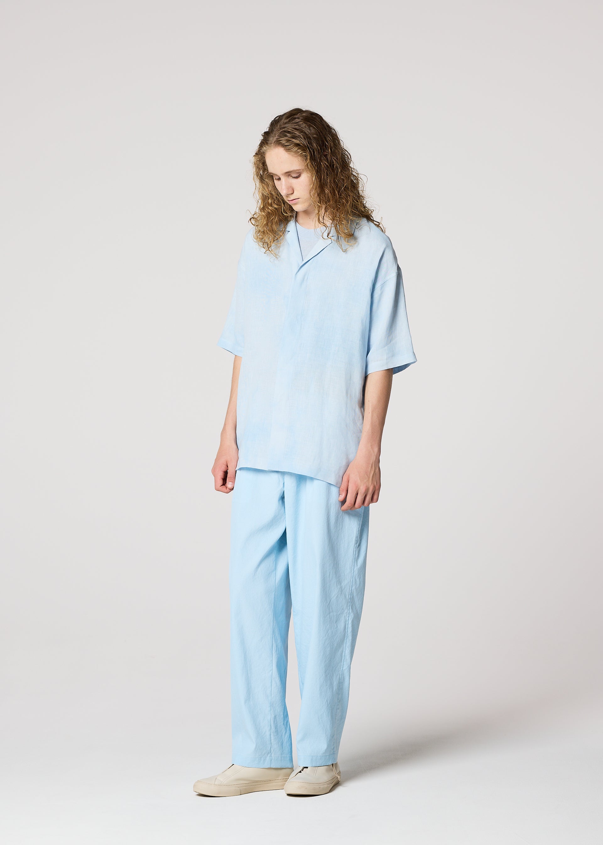 Zoomed view A person with long curly hair, dressed in IM MEN’s FLOATING LINEN loose-fitting shirt and matching pants, stands against a plain light background with a relaxed posture, looking down and wearing light-colored shoes.