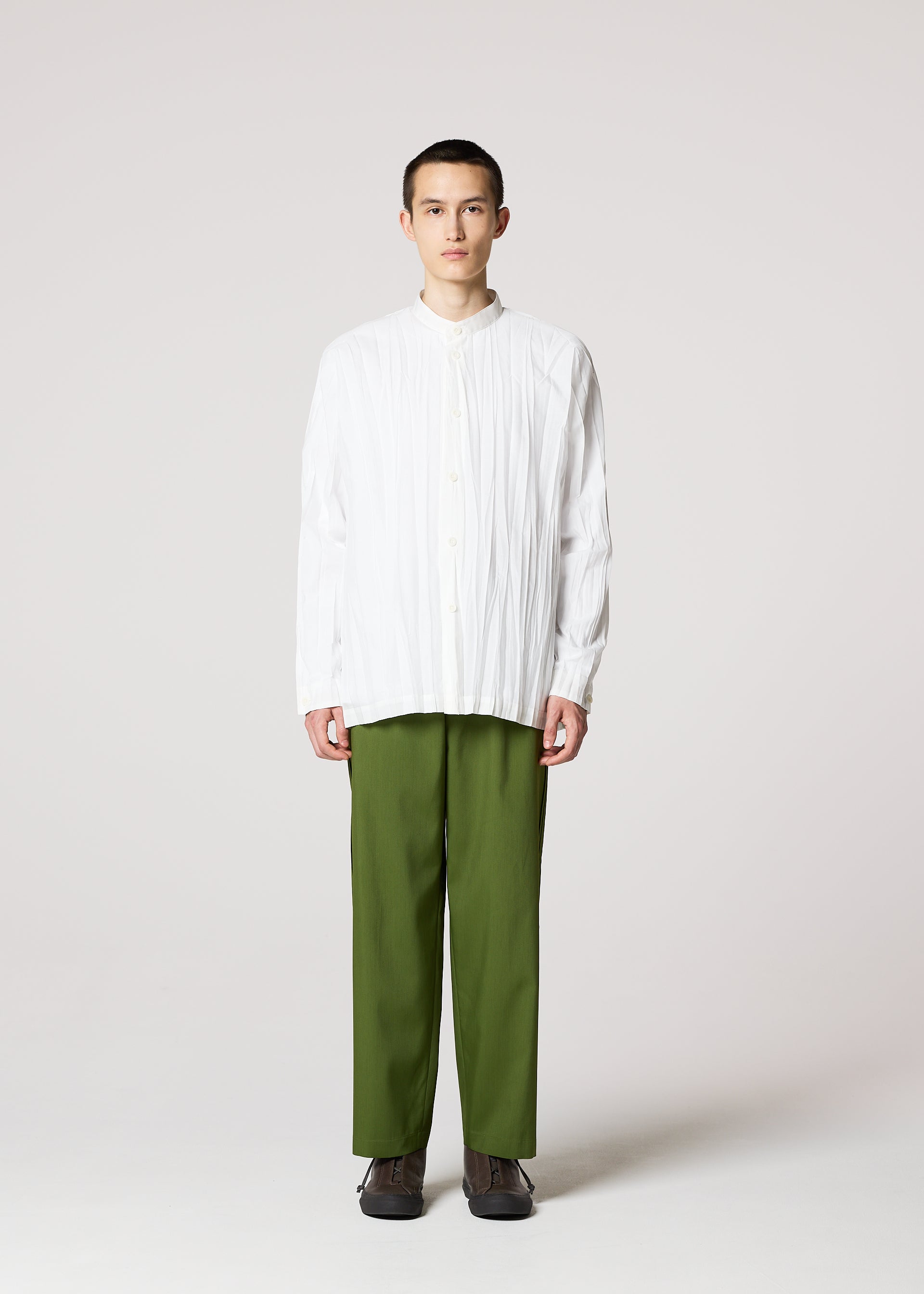 Zoomed view A person stands facing forward against a plain background, wearing the IM MEN WRINKLE SHIRT—a loose, long-sleeved white button-up with a unique creased finish—paired with green straight-leg pants and dark shoes.