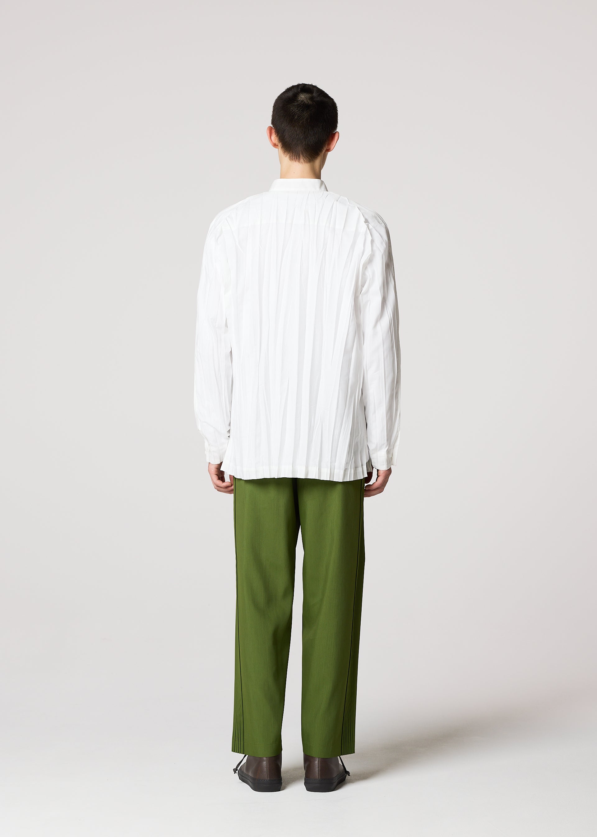Zoomed view A person faces away from the camera, wearing the IM MEN WRINKLE SHIRT made of recycled polyester with a unique creased finish, paired with olive green pants and dark shoes, set against a plain light gray background.