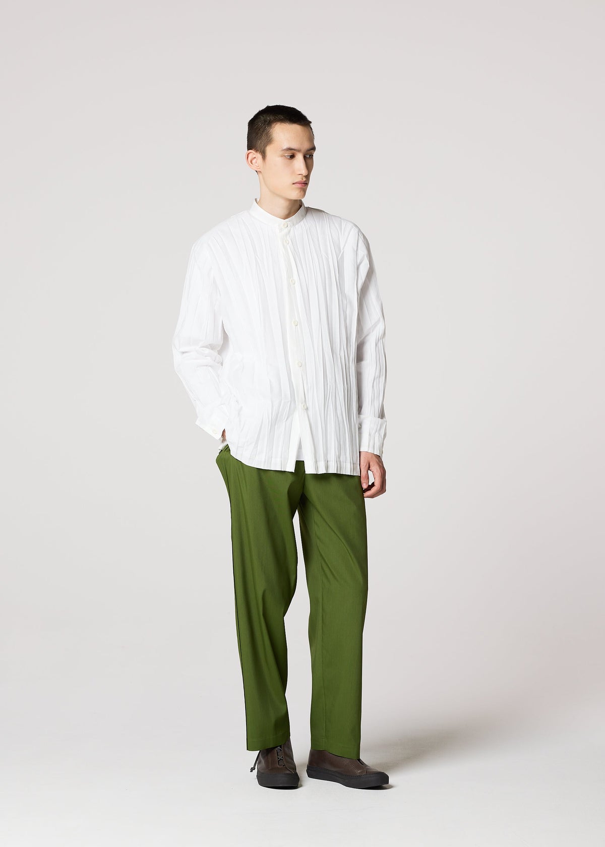 A young man with a neutral expression stands against a plain background, wearing the IM MEN WRINKLE SHIRT—a loose, long-sleeved shirt with a unique creased finish—paired with olive green pants and dark shoes, hands in his pockets. view 9 of 9