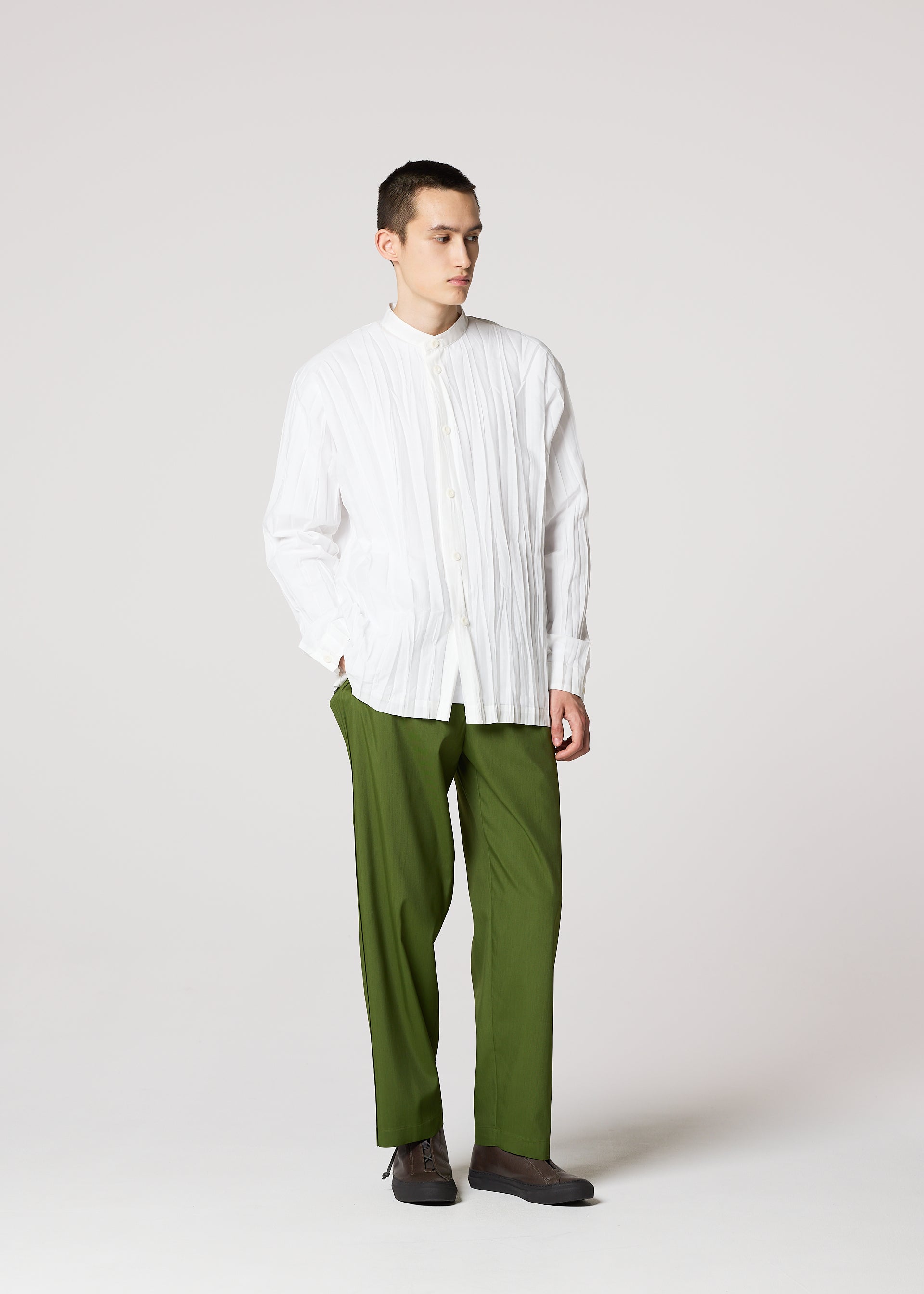Zoomed view A young man with a neutral expression stands against a plain background, wearing the IM MEN WRINKLE SHIRT—a loose, long-sleeved shirt with a unique creased finish—paired with olive green pants and dark shoes, hands in his pockets.