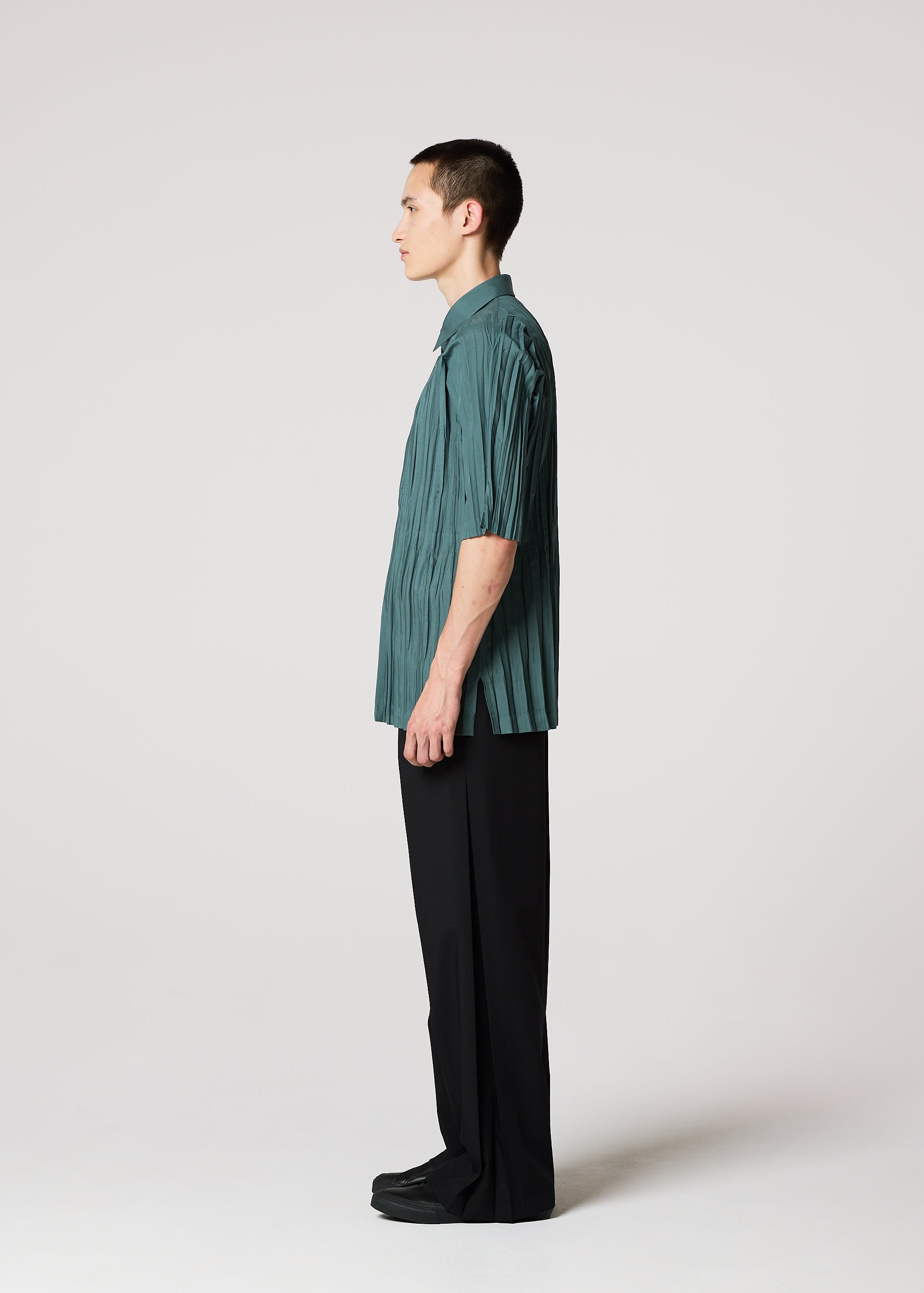 Zoomed view A person with short brown hair stands in profile against a white background, wearing the IM MEN WRINKLE SHIRT in teal recycled polyester, loose black pants, and black shoes.