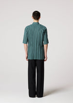 A person faces away from the camera, wearing the IM MEN WRINKLE SHIRT in teal recycled polyester with short sleeves and a pleated finish, paired with loose black pants against a plain light background.