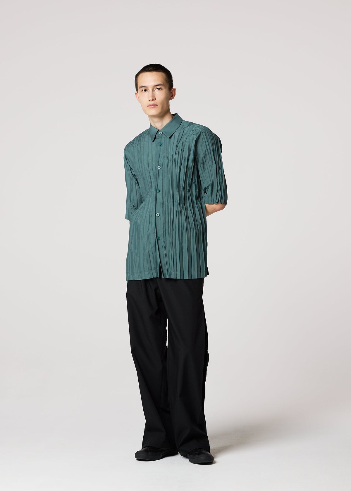 A person stands in a studio wearing the IM MEN WRINKLE SHIRT, a teal pleated recycled polyester short-sleeve, paired with loose black pants and black shoes against a plain light background. view 9 of 9