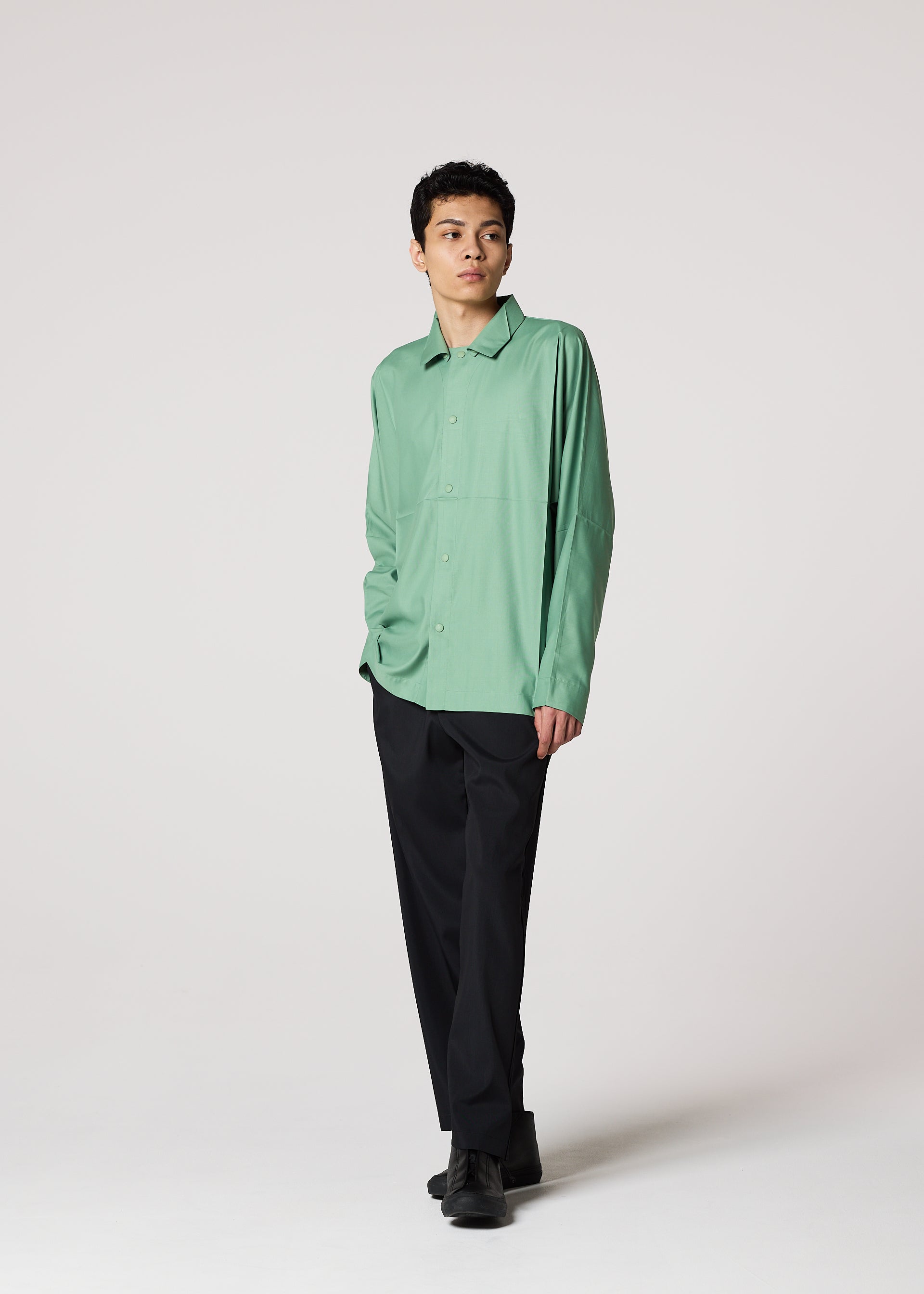 Zoomed view A person stands against a plain light background, wearing the IM MEN COMPACT SHIRT in light green, paired with black pants and black shoes, with one hand in their pocket and looking to the side.