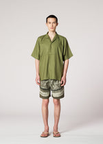 A person faces forward in an IM MEN COMPACT SHIRT (loose, wrinkle-resistant, olive green), landscape-print shorts, and sandals, set against a plain light background.