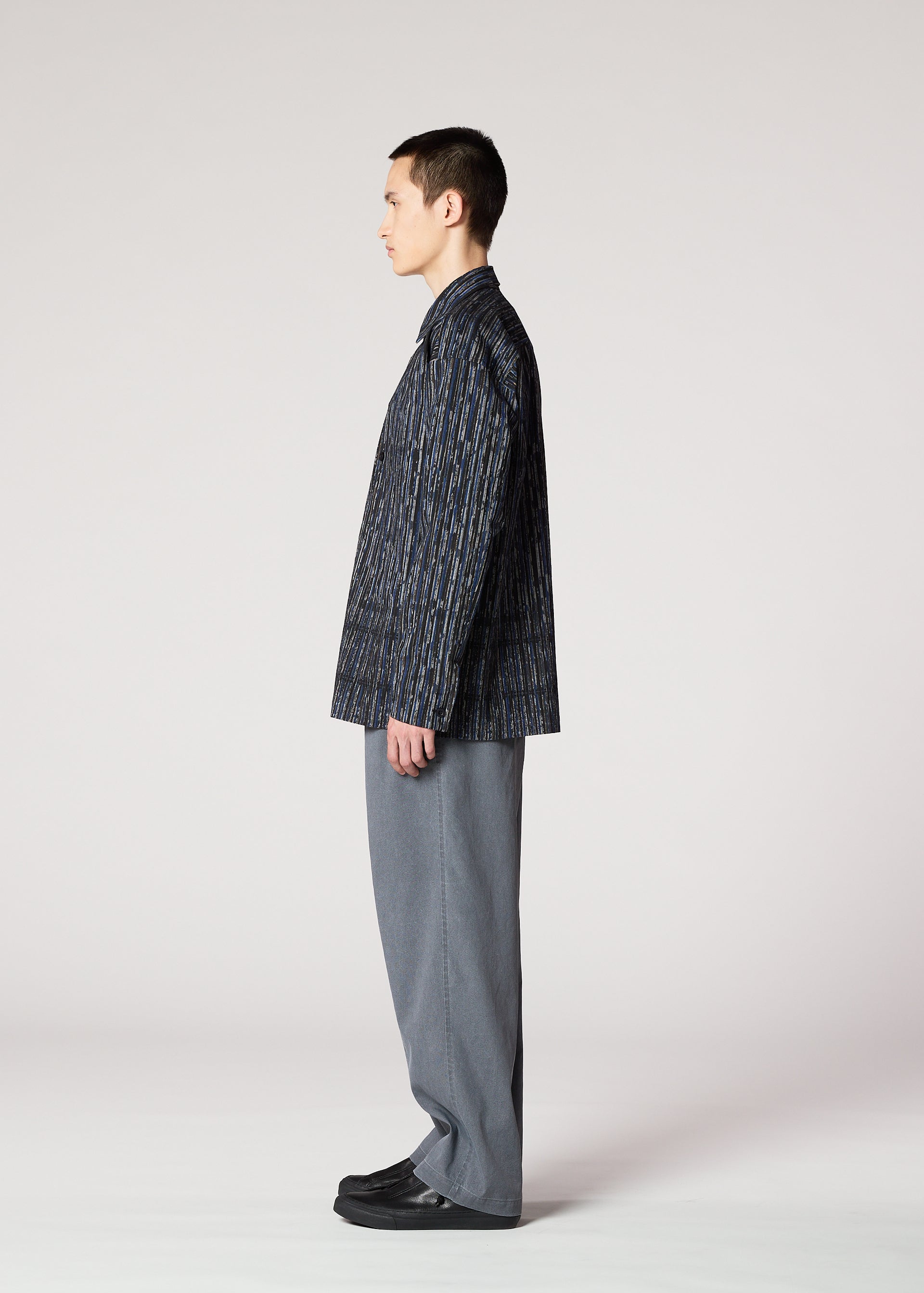 Zoomed view A person with short dark hair stands in profile against a plain background, wearing the IM MEN CURVINESS SHIRT, a dark patterned jacket, wide gray pants, and black shoes—evoking ceramic artist Shoji Kamoda's refined style.