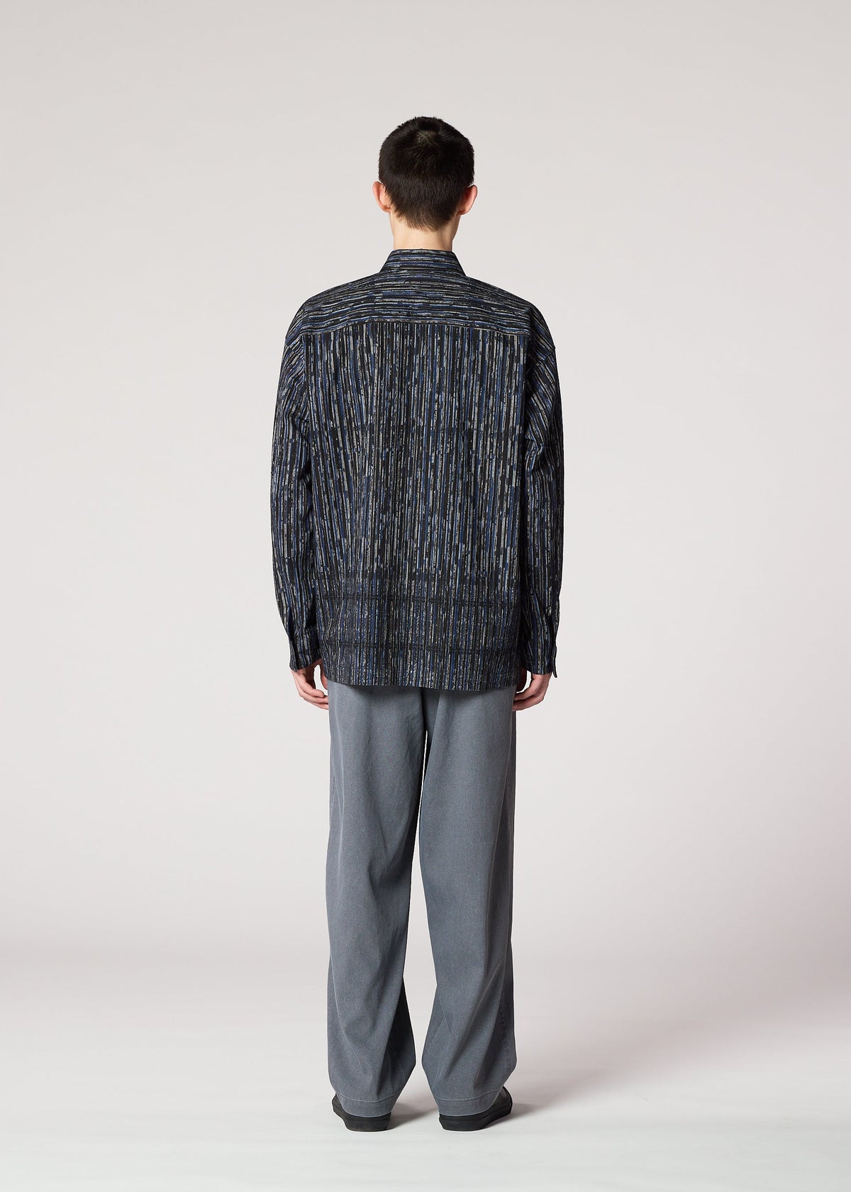 A person faces away from the camera, wearing the IM MEN CURVINESS SHIRT with a textured stripe and loose gray pants, echoing Shoji Kamoda’s minimalist style, set against a plain light background. view 7 of 8