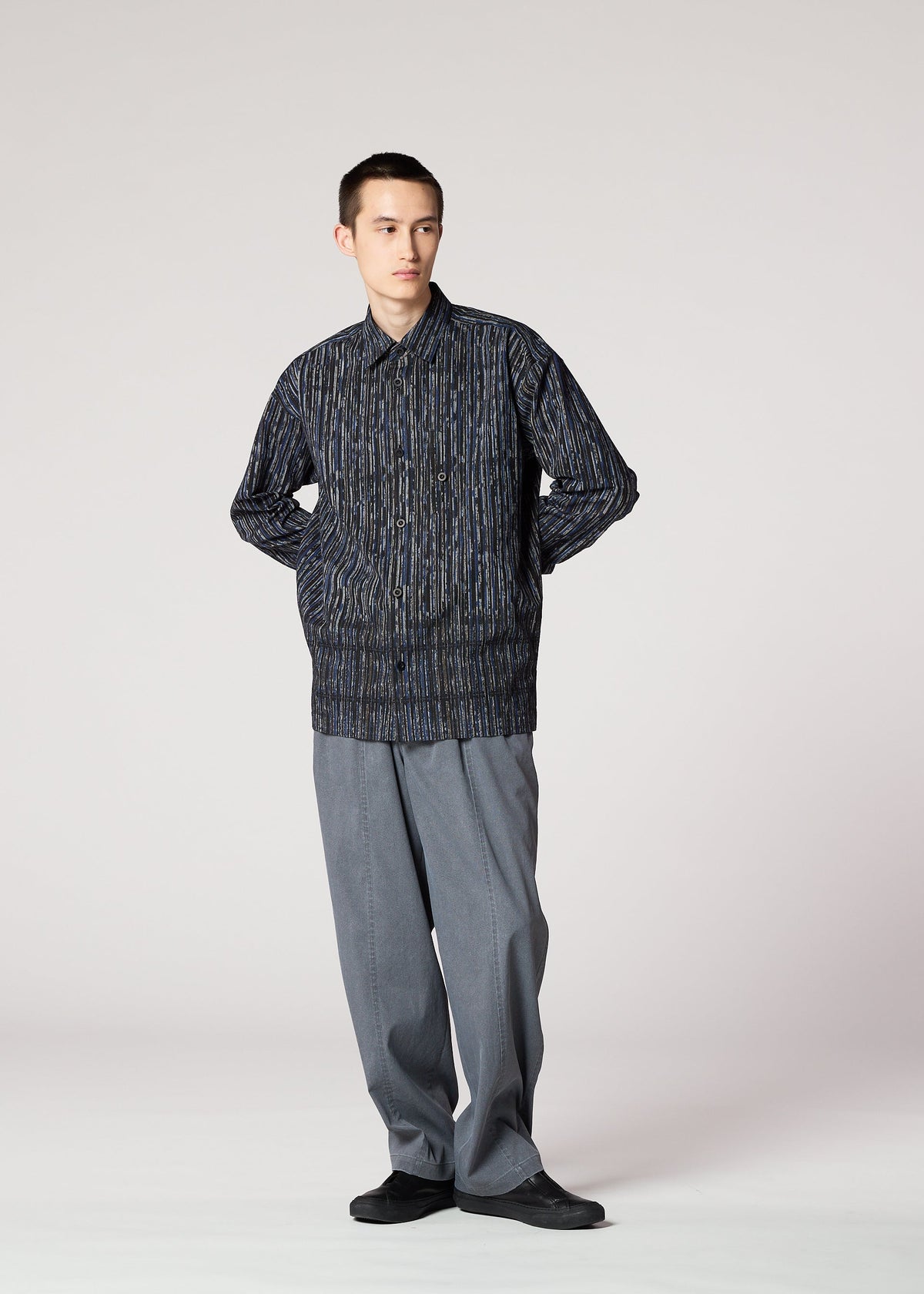 A person stands against a plain background, wearing the IM MEN CURVINESS SHIRT with textured stripes, loose gray trousers, and black shoes, hands in pockets, looking slightly to the side. view 8 of 8