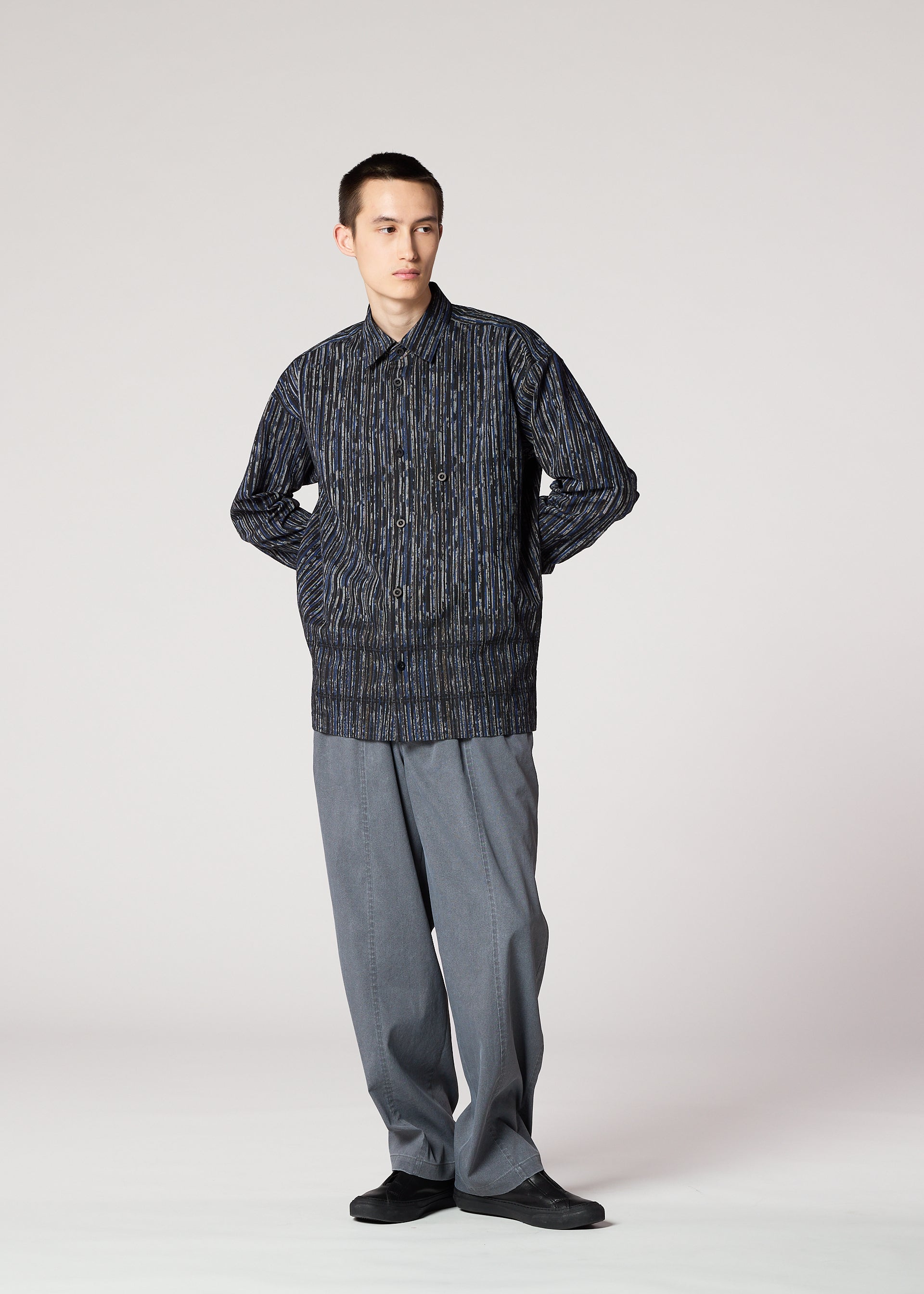 Zoomed view A person stands against a plain background, wearing the IM MEN CURVINESS SHIRT with textured stripes, loose gray trousers, and black shoes, hands in pockets, looking slightly to the side.