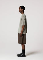 A person stands in profile against a plain light background, wearing the IM MEN SHRINK STRETCH T— a loose gray tee with uneven texture and deep colors—paired with brown knee-length shorts, black socks, and black shoes.