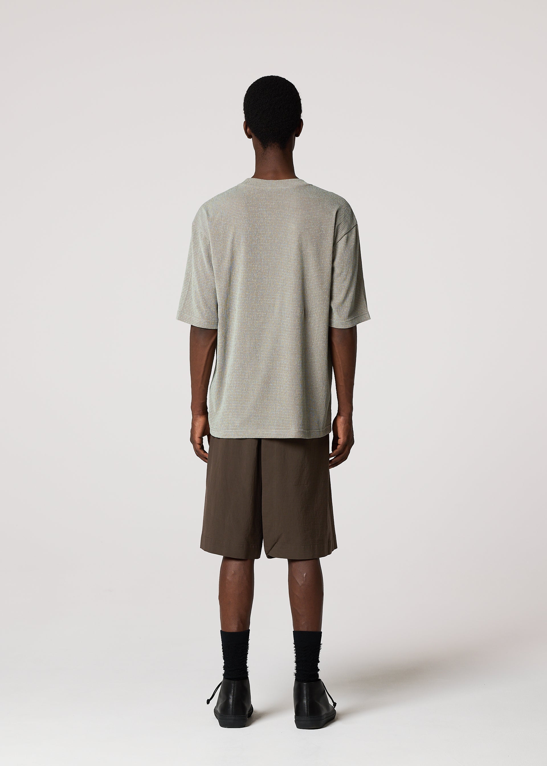 Zoomed view A person faces away from the camera, wearing the IM MEN SHRINK STRETCH T in grey, brown knee-length shorts, black socks, and black shoes against a plain light background.
