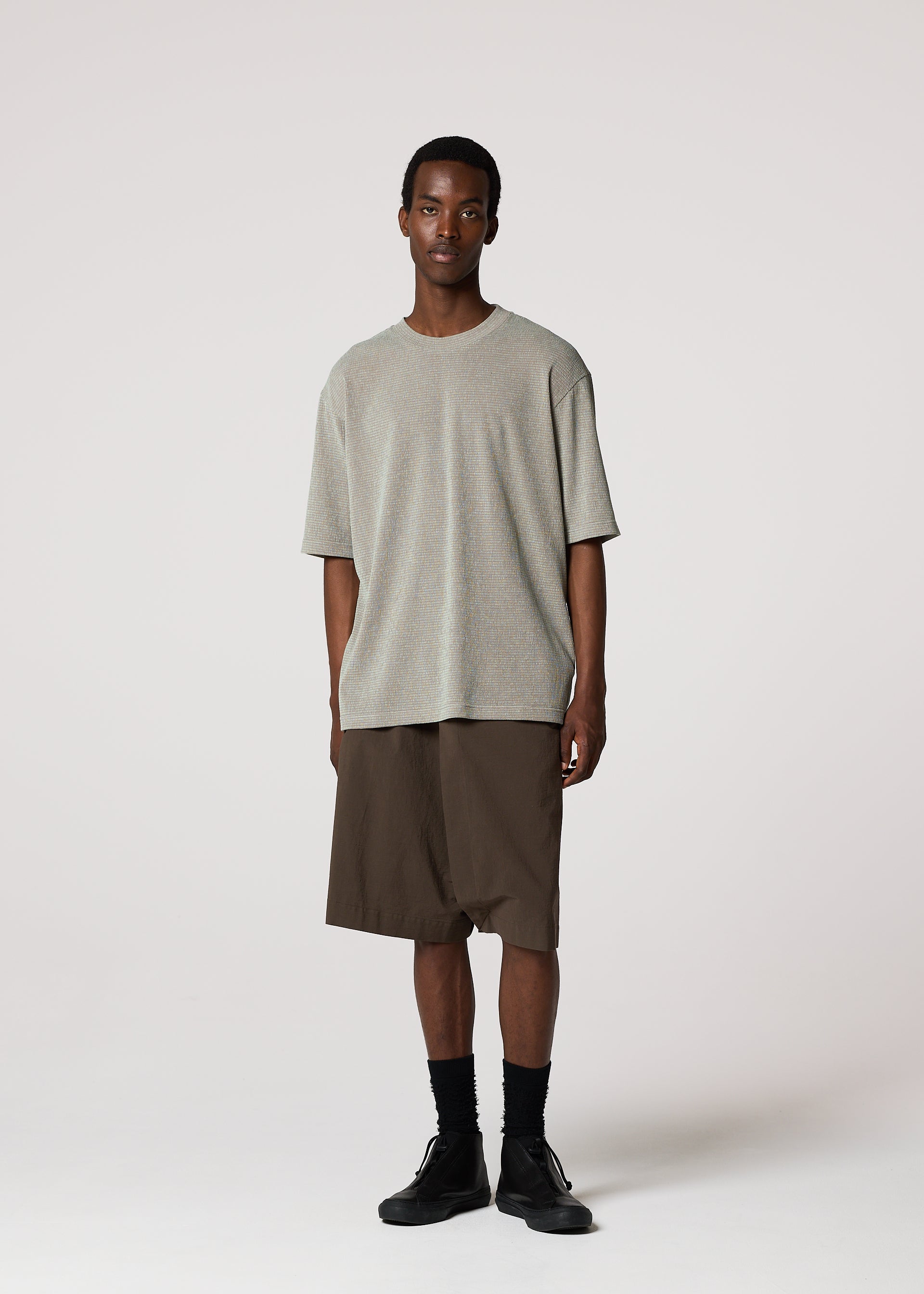 Zoomed view A man stands against a plain light background wearing the IM MEN SHRINK STRETCH T in gray, brown knee-length shorts, black socks, and black shoes. He faces forward with a neutral expression and relaxed posture.
