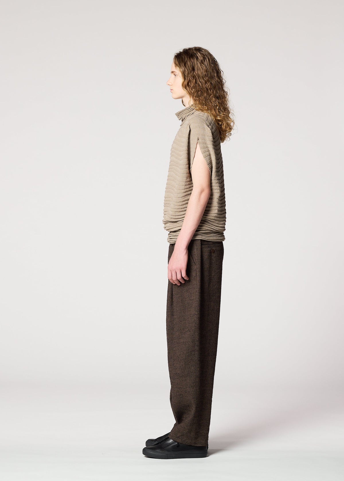 A person with long curly hair stands in profile against a plain light background, wearing the IM MEN ENGRAVE KNIT top with wide, dark brown pants and black shoes. view 6 of 8