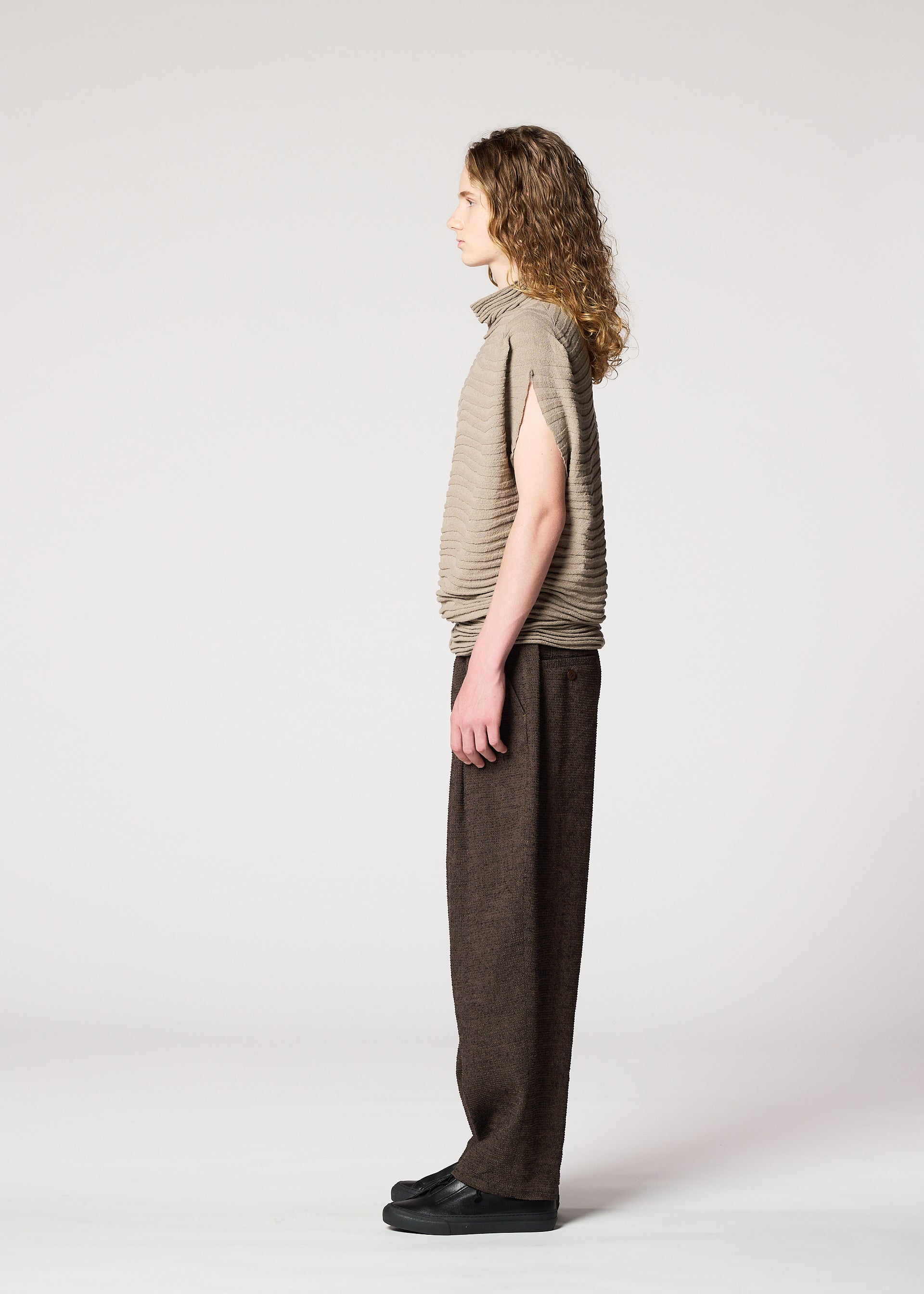 Zoomed view A person with long curly hair stands in profile against a plain light background, wearing the IM MEN ENGRAVE KNIT top with wide, dark brown pants and black shoes.