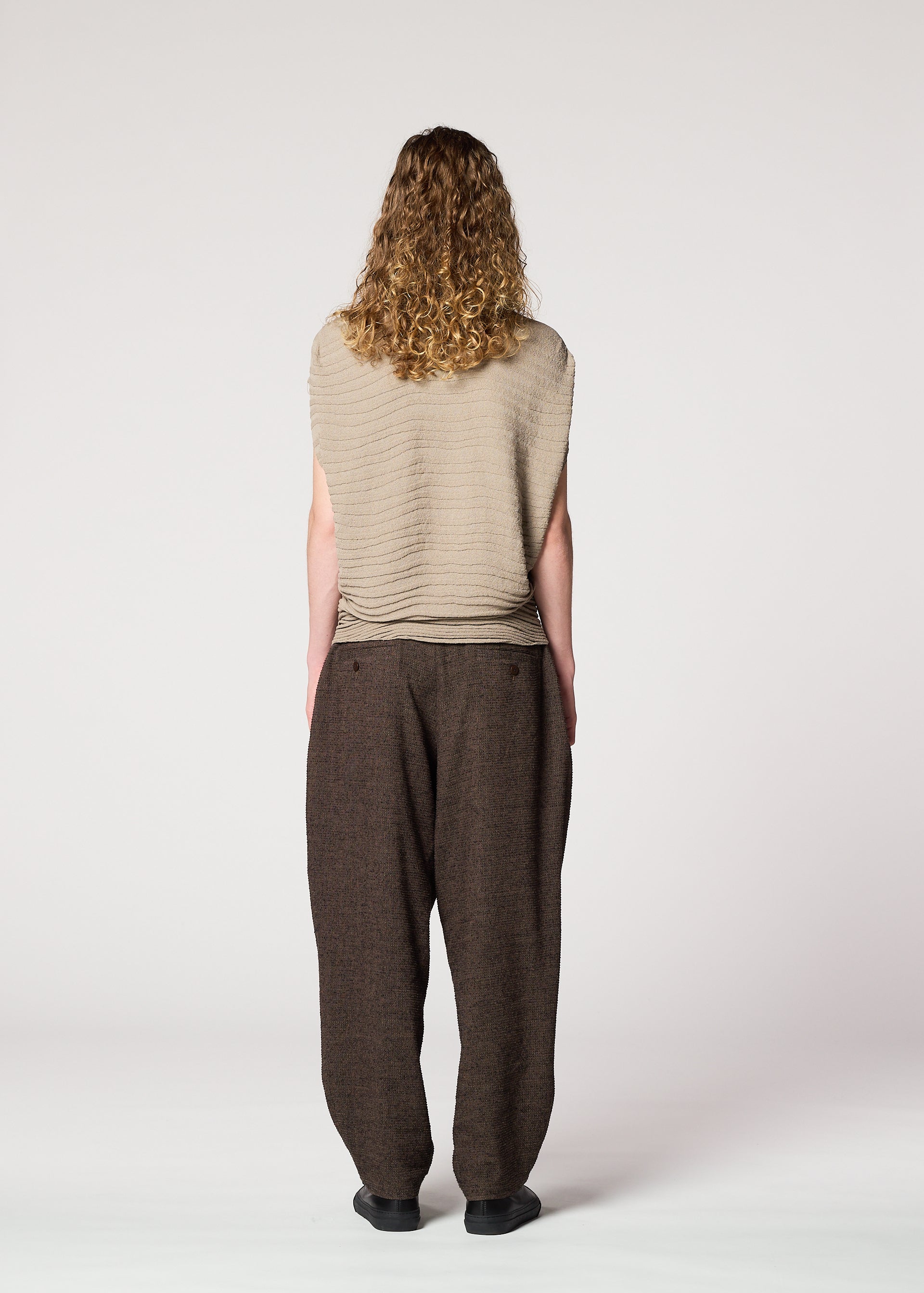 Zoomed view A person with long curly hair stands facing away, wearing the IM MEN ENGRAVE KNIT top and baggy brown pants with black shoes, against a plain light-colored background.