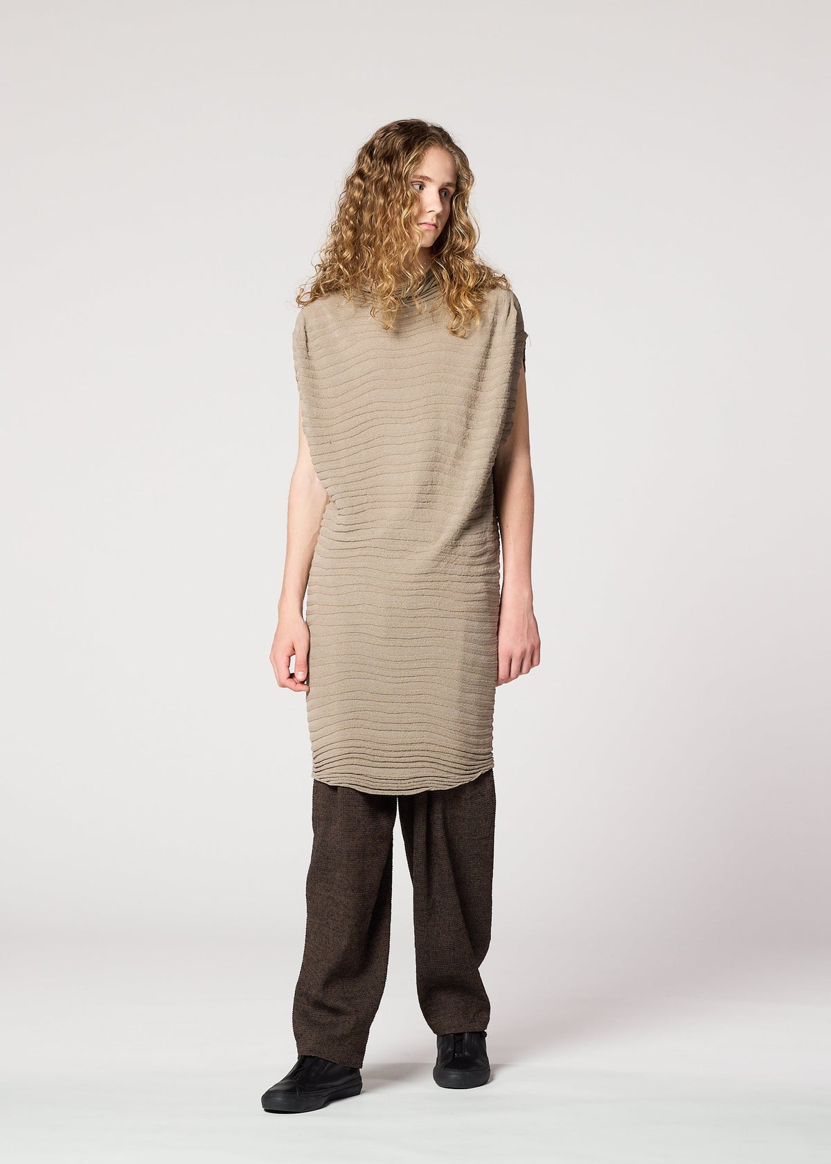A person with long curly hair wears IM MEN’s ENGRAVE KNIT in beige, paired with wide-legged brown pants and black shoes, inspired by Shoji Kamoda, standing against a plain light background. view 8 of 8