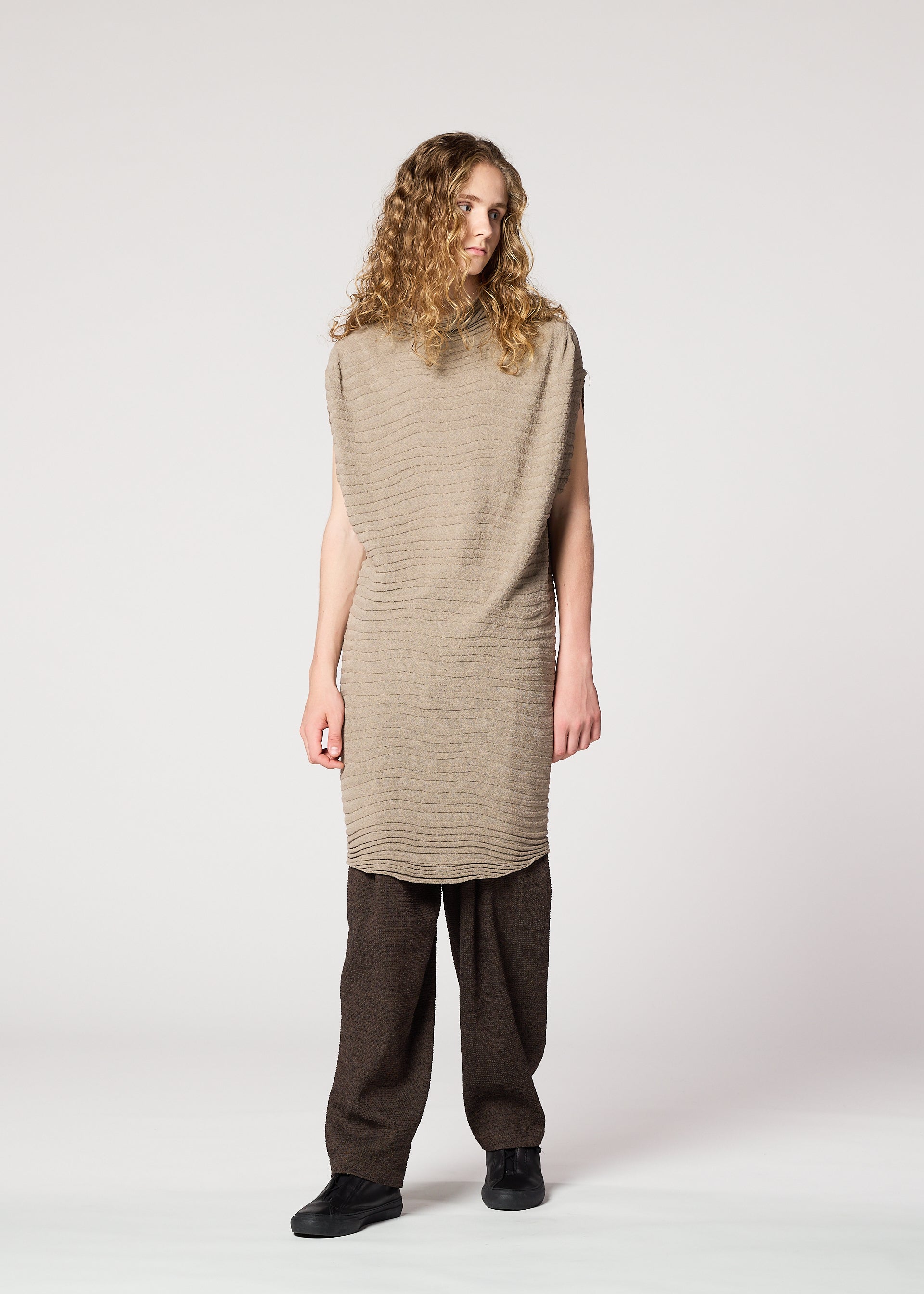 Zoomed view A person with long curly hair wears IM MEN’s ENGRAVE KNIT in beige, paired with wide-legged brown pants and black shoes, inspired by Shoji Kamoda, standing against a plain light background.