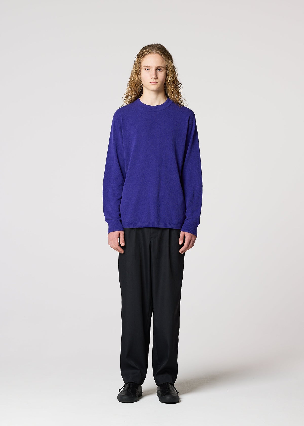 A person with long curly hair wears the IM MEN COMFORT loose, breathable, long-sleeved purple knit sweater made of cotton nylon yarn, paired with black pants and shoes, standing facing forward against a plain light background. view 5 of 8