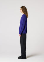 A person with long curly hair wears the IM MEN COMFORT purple long-sleeved knit top, made from breathable cotton nylon yarn, along with black pants and black shoes, standing in profile against a plain white background.