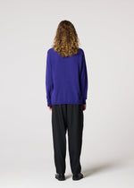 A person with curly, shoulder-length hair wears the IM MEN COMFORT purple knit long-sleeved sweater, black pants, and black shoes, standing with their back to a plain light background.