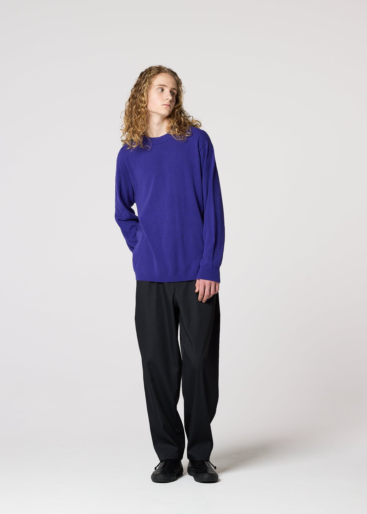 Against a plain background, a person with long curly hair wears the IM MEN COMFORT breathable, long-sleeved knit purple sweater, paired with black pants and shoes. They stand relaxed, looking slightly to the side. view 8 of 8
