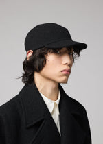 Person with medium-length dark hair wears the IM MEN FULLER WOOL CAP in black, paired with a black coat and white collared shirt, gazing slightly to the side against a plain gray background.