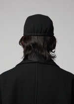 A person with medium-length dark hair, seen from behind in a black coat and the IM MEN FULLER WOOL CAP, stands against a plain light background.