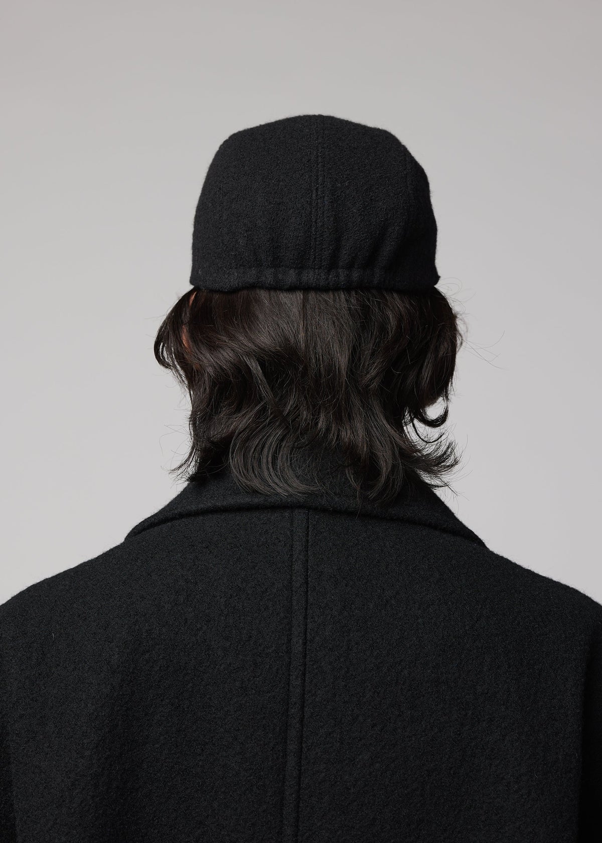 Zoomed view A person with medium-length dark hair, seen from behind in a black coat and the IM MEN FULLER WOOL CAP, stands against a plain light background.