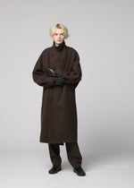 A person with short blond hair wears a long dark brown overcoat, brown pants, IM MEN PIGEON GLOVES in natural leather, and black shoes, standing against a plain light gray background.