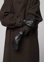 A person in a long, dark brown coat wears IM MEN PIGEON GLOVES, arms crossed and adjusting one glove; the image is cropped to show only their torso and hands.