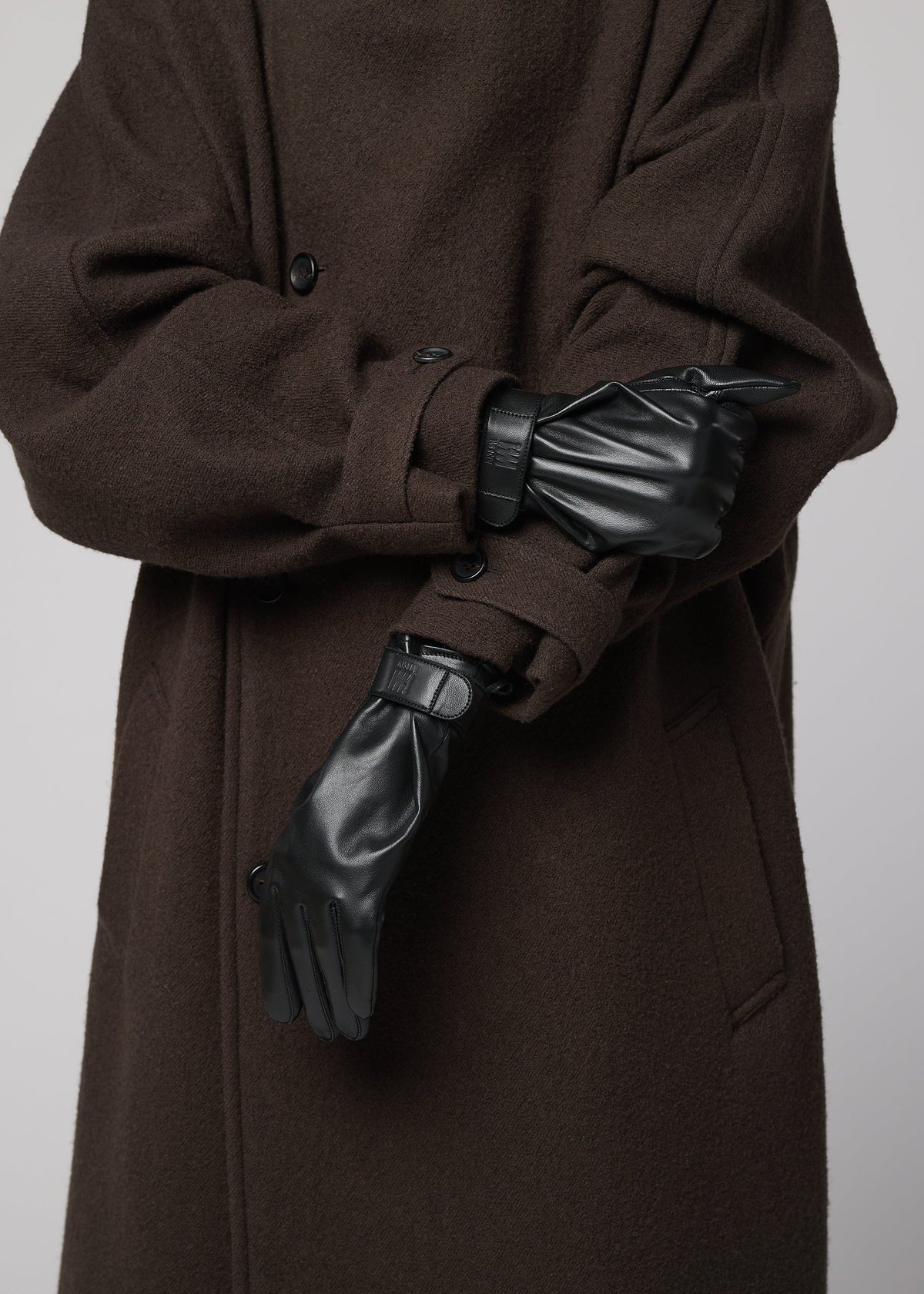 Zoomed view A person in a long, dark brown coat wears IM MEN PIGEON GLOVES, arms crossed and adjusting one glove; the image is cropped to show only their torso and hands.