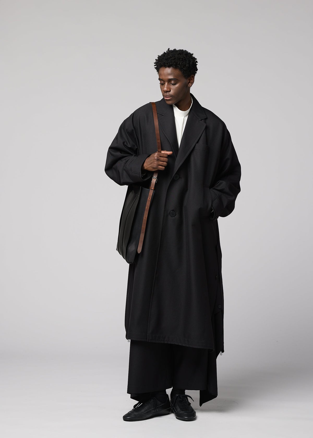 A person wearing a long, oversized black coat, black pants, and black shoes stands against a plain gray background, looking down and holding the IM MEN LEATHER PLEATS shoulder bag with a natural leather body and brown strap. view 7 of 8