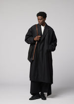 A person wearing a long, oversized black coat, black pants, and black shoes stands against a plain gray background, looking down and holding the IM MEN LEATHER PLEATS shoulder bag with a natural leather body and brown strap.