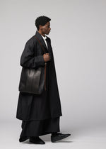 An individual dressed in a long black coat and wide black pants from IM MEN walks sideways, carrying the LEATHER PLEATS large pleated shoulder bag. They pair the look with black shoes and a white turtleneck, set against a plain light gray background.