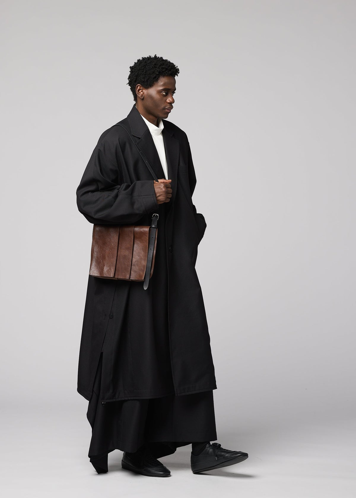A person dressed in a long black coat, black pants, and black shoes is holding the IM MEN LEATHER PLEATS natural-leather shoulder bag. The plain light gray background echoes the sophisticated aesthetic of the IM MEN Autumn Winter 2026/27 Collection. view 7 of 8