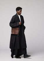 A person dressed in a long black coat, black pants, and black shoes is holding the IM MEN LEATHER PLEATS natural-leather shoulder bag. The plain light gray background echoes the sophisticated aesthetic of the IM MEN Autumn Winter 2026/27 Collection.