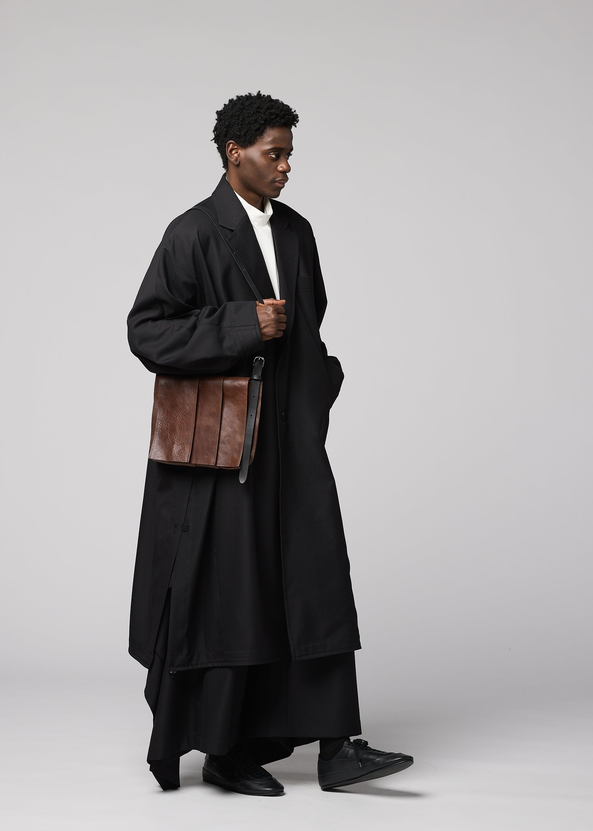 Zoomed view A person dressed in a long black coat, black pants, and black shoes is holding the IM MEN LEATHER PLEATS natural-leather shoulder bag. The plain light gray background echoes the sophisticated aesthetic of the IM MEN Autumn Winter 2026/27 Collection.