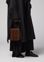 A person dressed in a long, black coat and black shoes holds the LEATHER PLEATS brown natural-leather shoulder bag by their side. The image—cropped to display only the lower half of the body against a plain background—captures a sleek look from IM MEN’s Autumn Winter 2026/27 Collection.