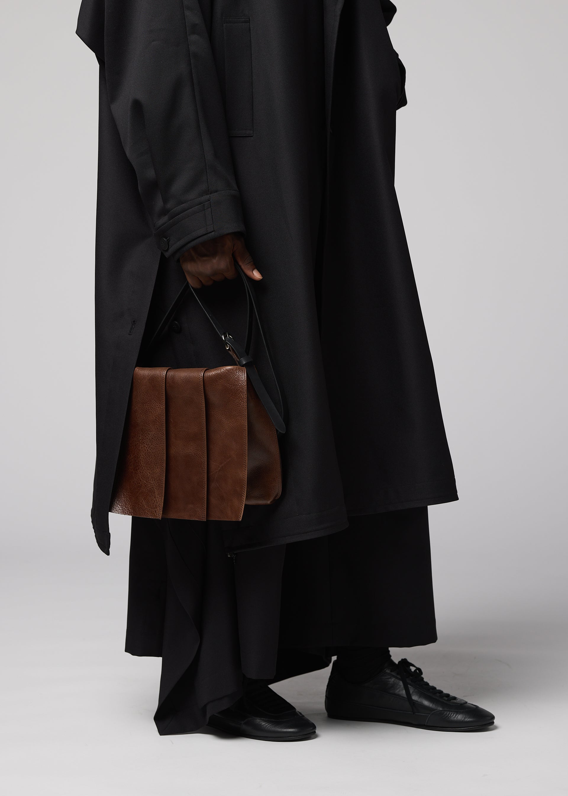 Zoomed view A person dressed in a long, black coat and black shoes holds the LEATHER PLEATS brown natural-leather shoulder bag by their side. The image—cropped to display only the lower half of the body against a plain background—captures a sleek look from IM MEN’s Autumn Winter 2026/27 Collection.
