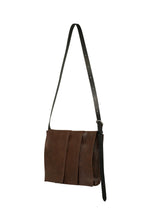 The LEATHER PLEATS shoulder bag by IM MEN, from the Autumn Winter 2026/27 Collection, is crafted in natural brown leather with a long, adjustable black strap and features a layered front flap design reminiscent of pleats. Shown against a white background.