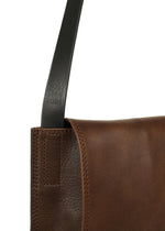 Close-up of the LEATHER PLEATS shoulder bag by IM MEN, crafted from brown natural leather with visible stitching and a black strap. The strap, part of the IM MEN Autumn Winter 2026/27 Collection, features embossed text near the edge of the bag.