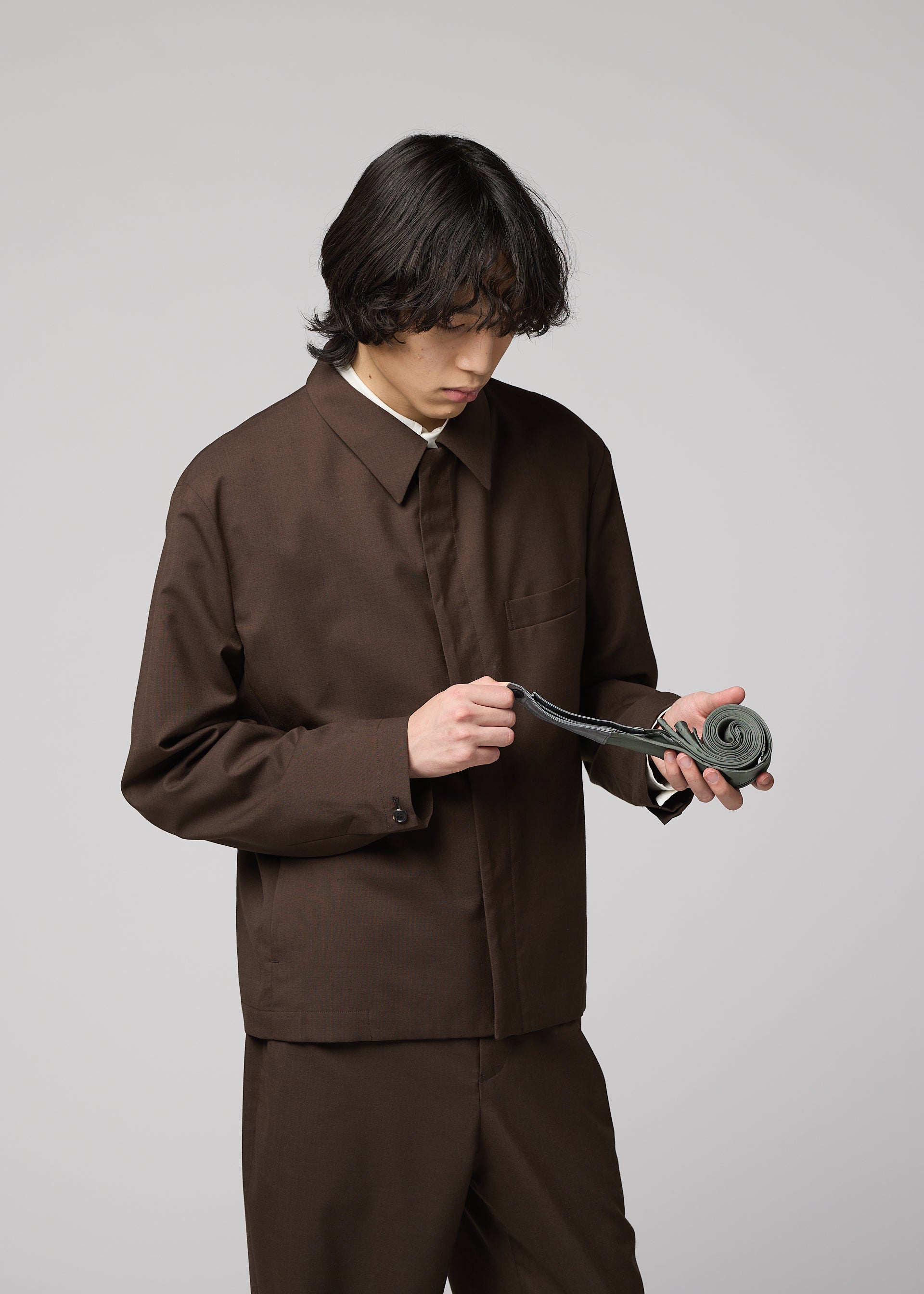 Zoomed view A person with medium-length dark hair in a brown suit looks down while unrolling the handmade-pleat gray GURU GURU tie by IM MEN against a plain light background.