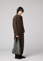 A person with medium-length dark hair stands against a plain light background, wearing a loose brown suit and black shoes, holding the light green GURU GURU foldable tote bag with handmade pleats by IM MEN in their left hand.